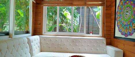 A room with a white cushioned bench, a small table with a decorative bowl, and a large window with a view of greenery.