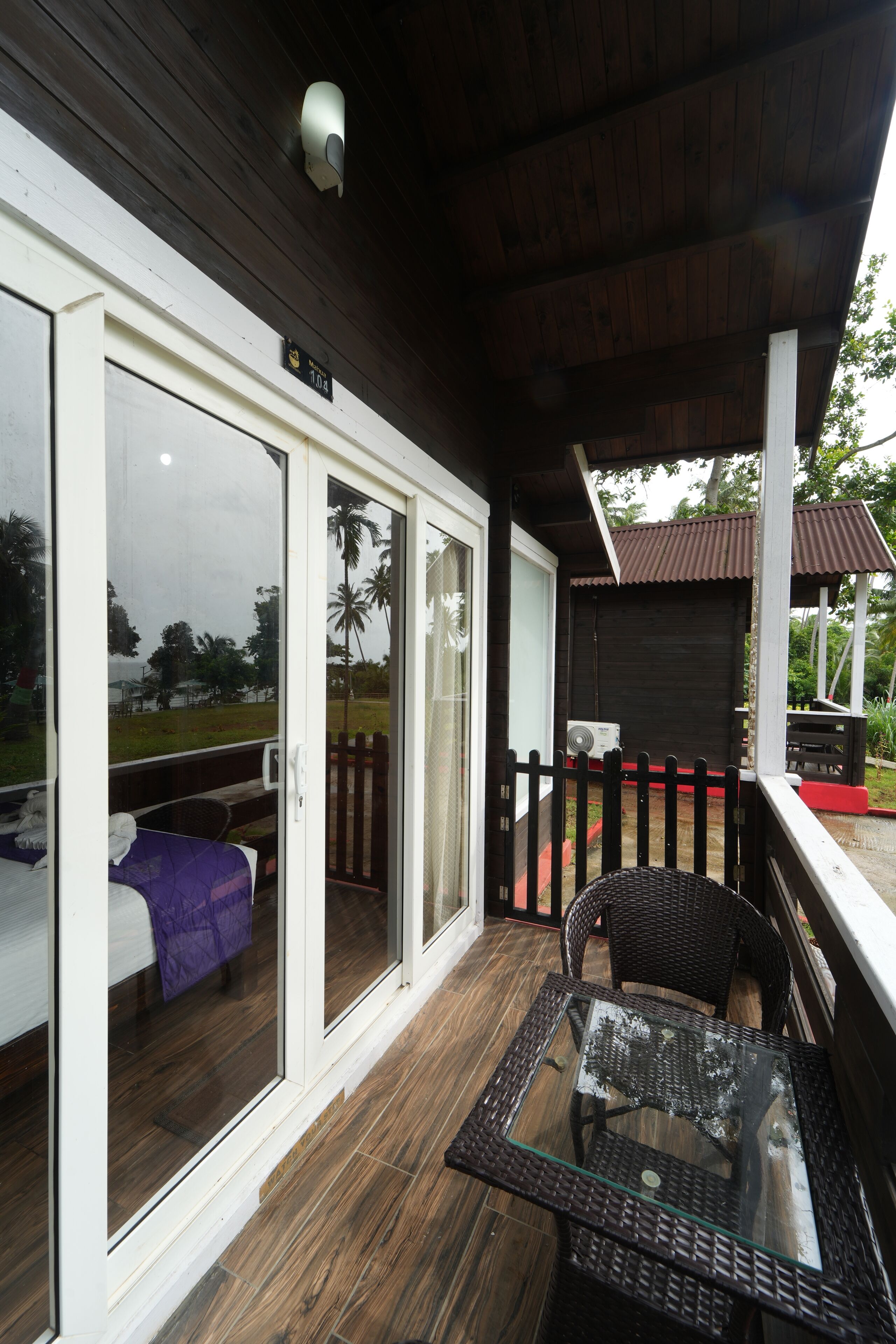 Un espacio exterior cubierto con una puerta de vidrio que da a una habitación, una mesa de vidrio y una terraza de madera.