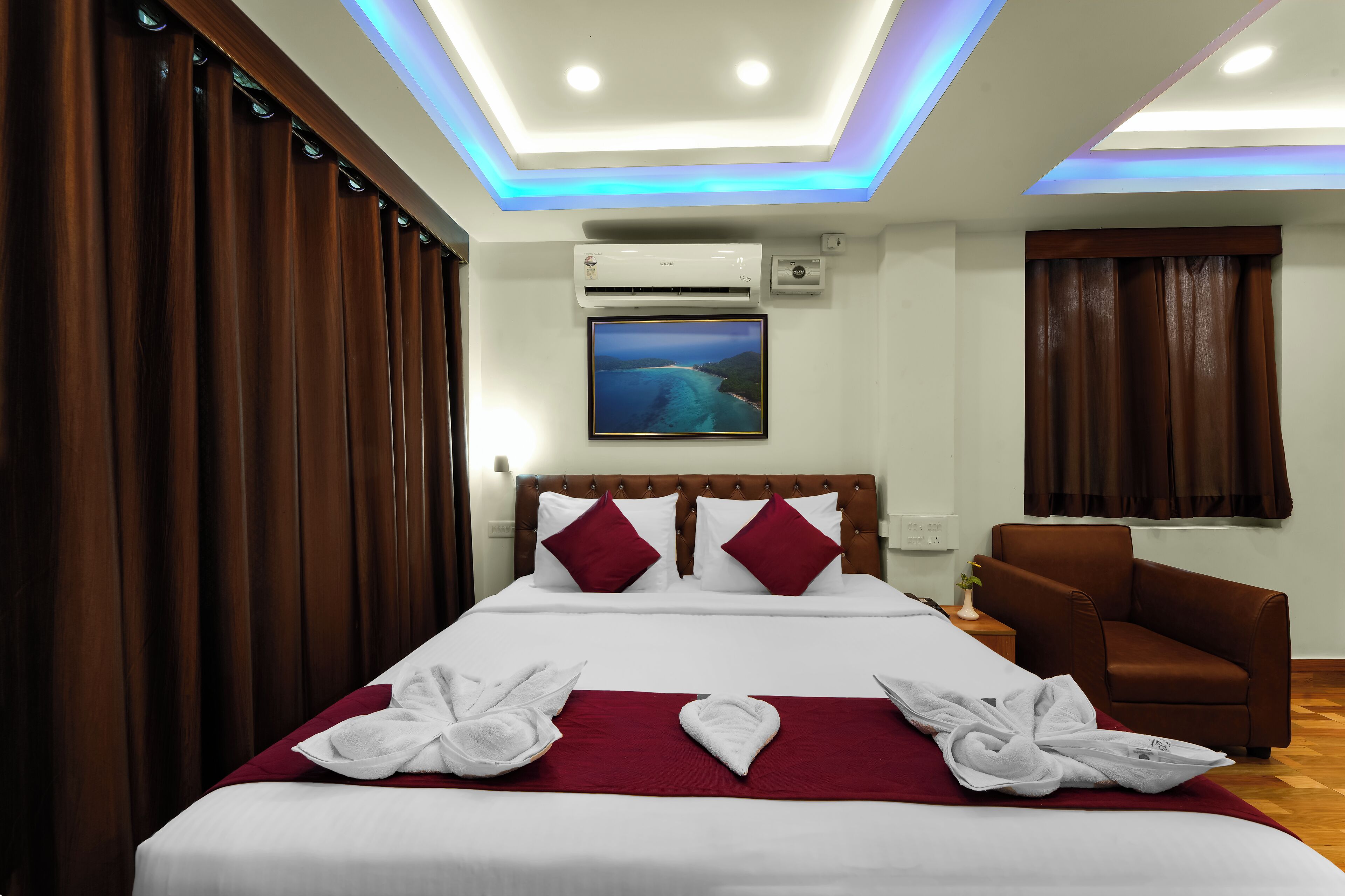 Une chambre d’hôtel avec un grand lit, un fauteuil en cuir marron, un appareil de climatisation, un téléviseur fixé au mur et une fenêtre avec des rideaux.