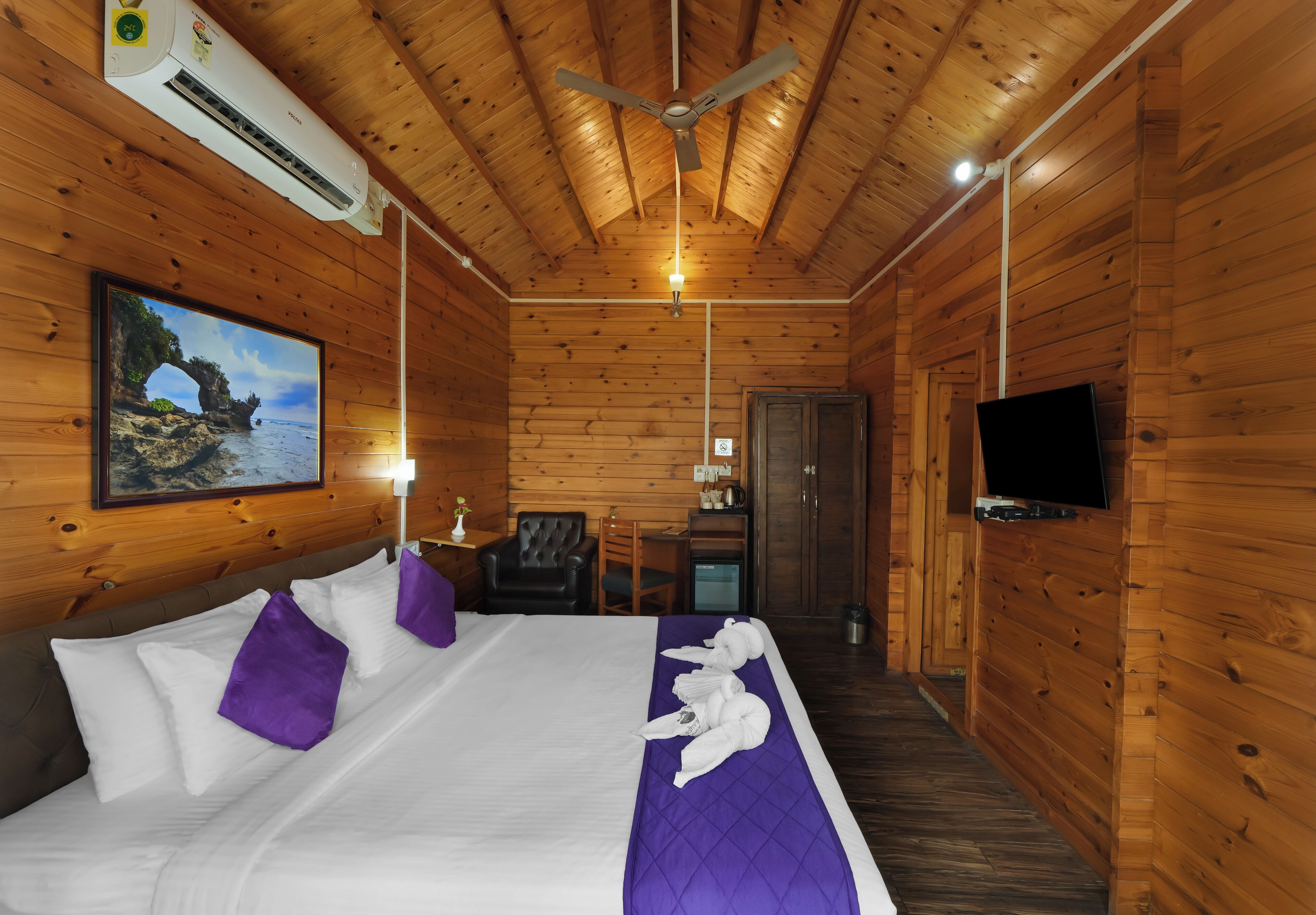 Une chambre dans un chalet en bois, équipée d’un lit, d’un ventilateur de plafond, d’une télévision et ornée d’un tableau représentant un paysage côtier.
