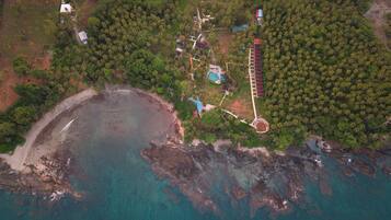 An aerial view of a coastal property with a beach, clear waters, dense palm trees, and a swimming pool.