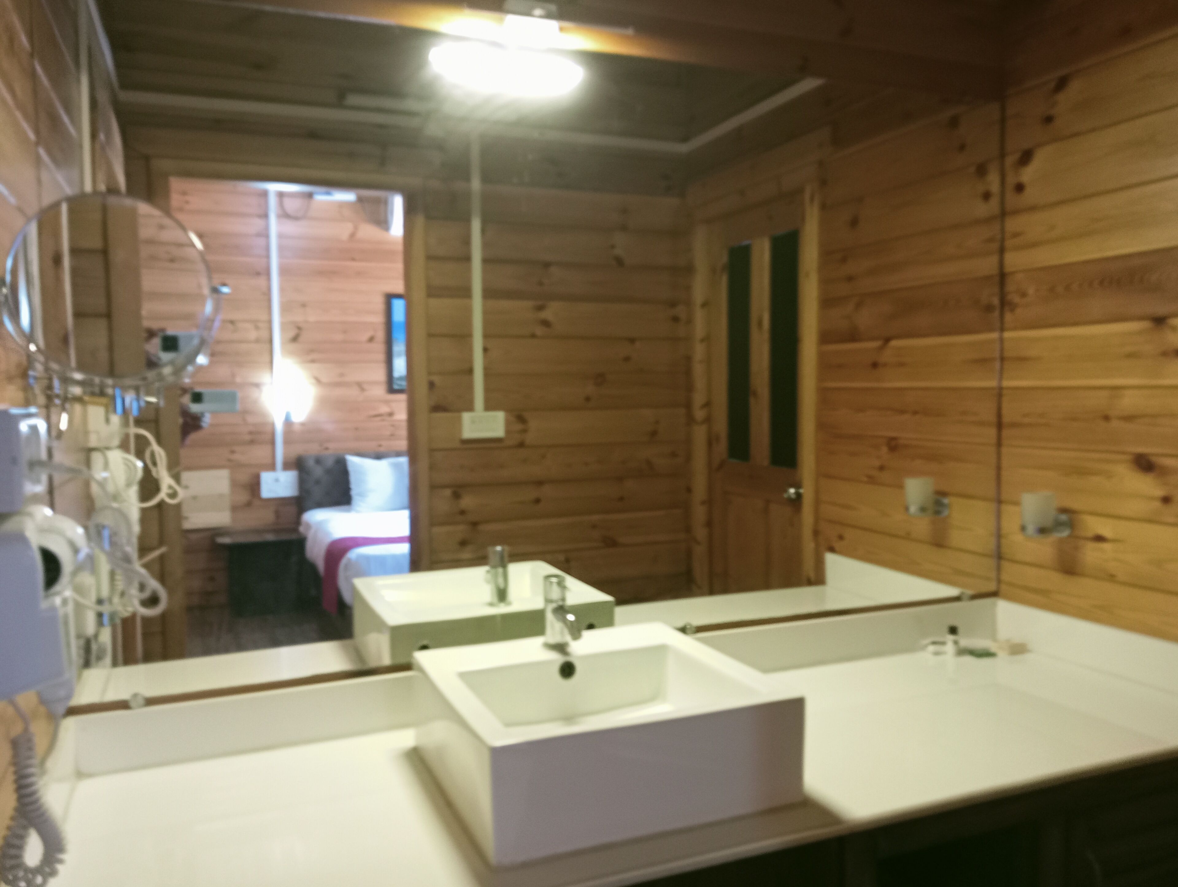 A wooden cabin interior with a bathroom featuring two sinks, a mirror, and a bed visible through an open door.