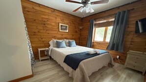 Een slaapkamer met een bed, nachtkastje, plafondventilator, raam met gordijnen en een televisie aan de muur.