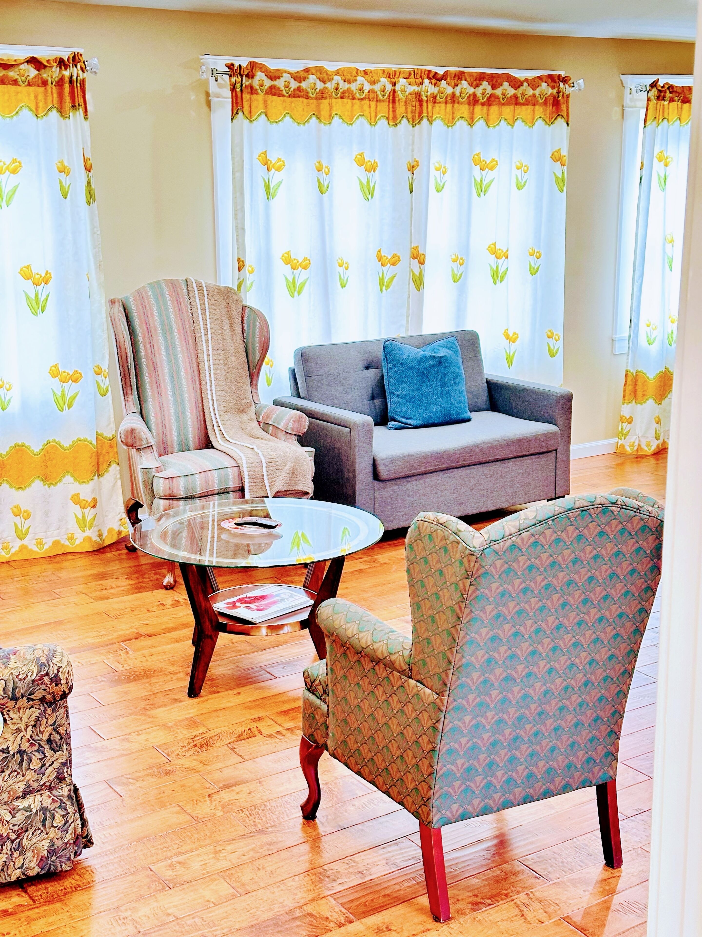 A living room with a striped armchair, a patterned armchair, a grey sofa with a blue pillow, and a glass coffee table.
