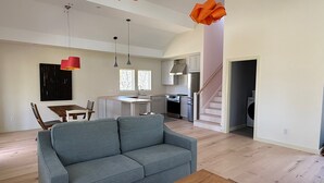 A spacious living area with a grey sofa, wooden coffee table, and a dining table with chairs. A kitchen with modern appliances and a staircase leading to another level is visible in the background. - Newly Renovated Charming Guest House in Chilmark with WiFi, AC, Covered Porch (Chilmark)