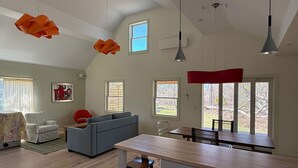 A spacious living area with a sofa, a dining table, and a kitchen island. There are multiple windows providing natural light and views of the outdoors. - Newly Renovated Charming Guest House in Chilmark with WiFi, AC, Covered Porch (Chilmark)