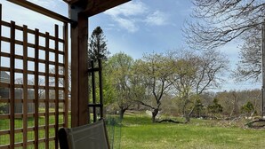 A covered patio area with a wooden fence, a chair, and a view of a grassy landscape with trees. - Newly Renovated Charming Guest House in Chilmark with WiFi, AC, Covered Porch (Chilmark)