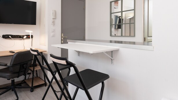 Ein modernes Zimmer mit Schreibtisch, zwei schwarzen Stühlen, einem Wand-TV, einer Tür und einem Spiegel.