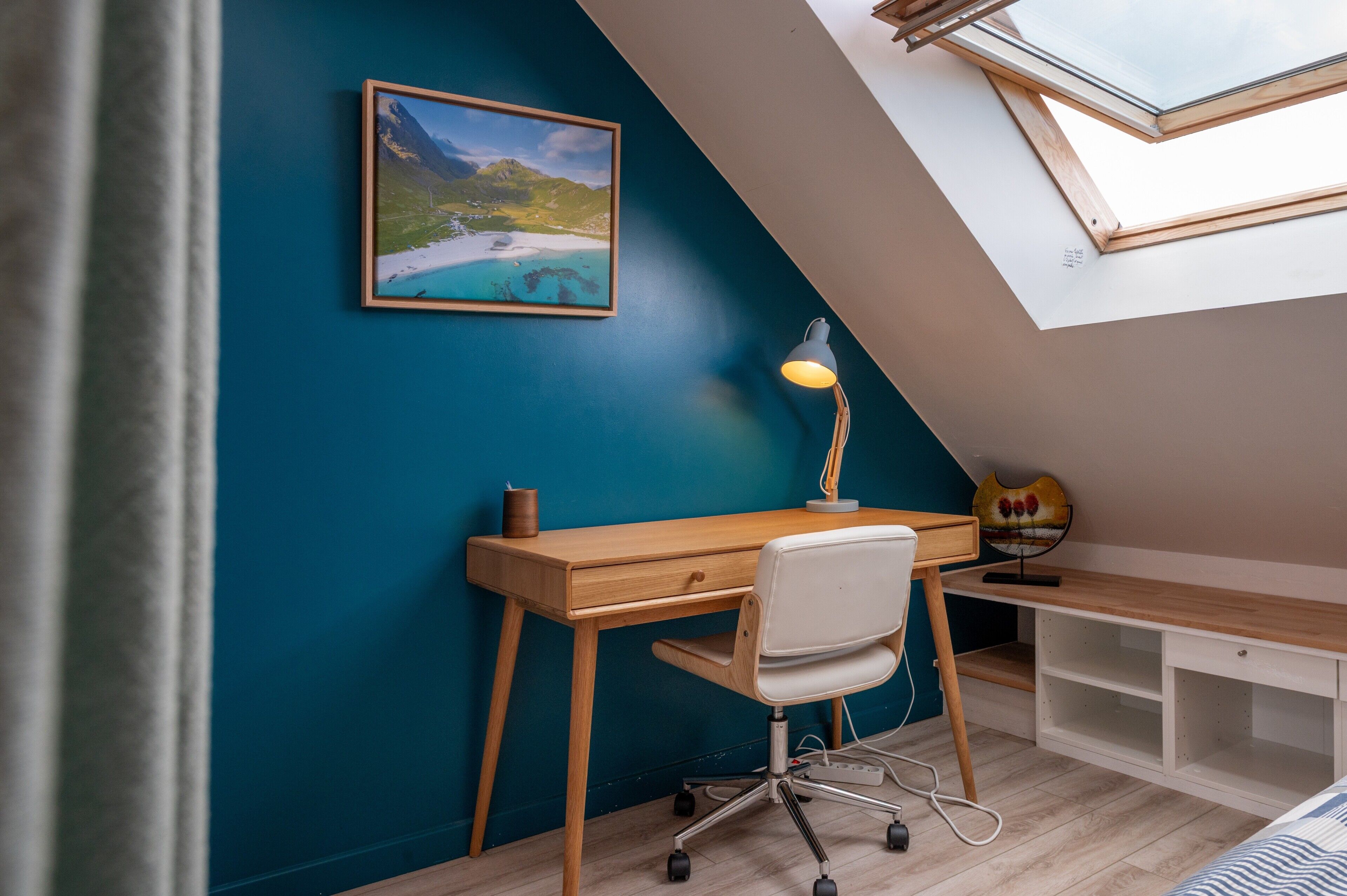 A room with a sloped ceiling, a desk with a chair, a lamp, a shelf, and a framed picture on the wall.