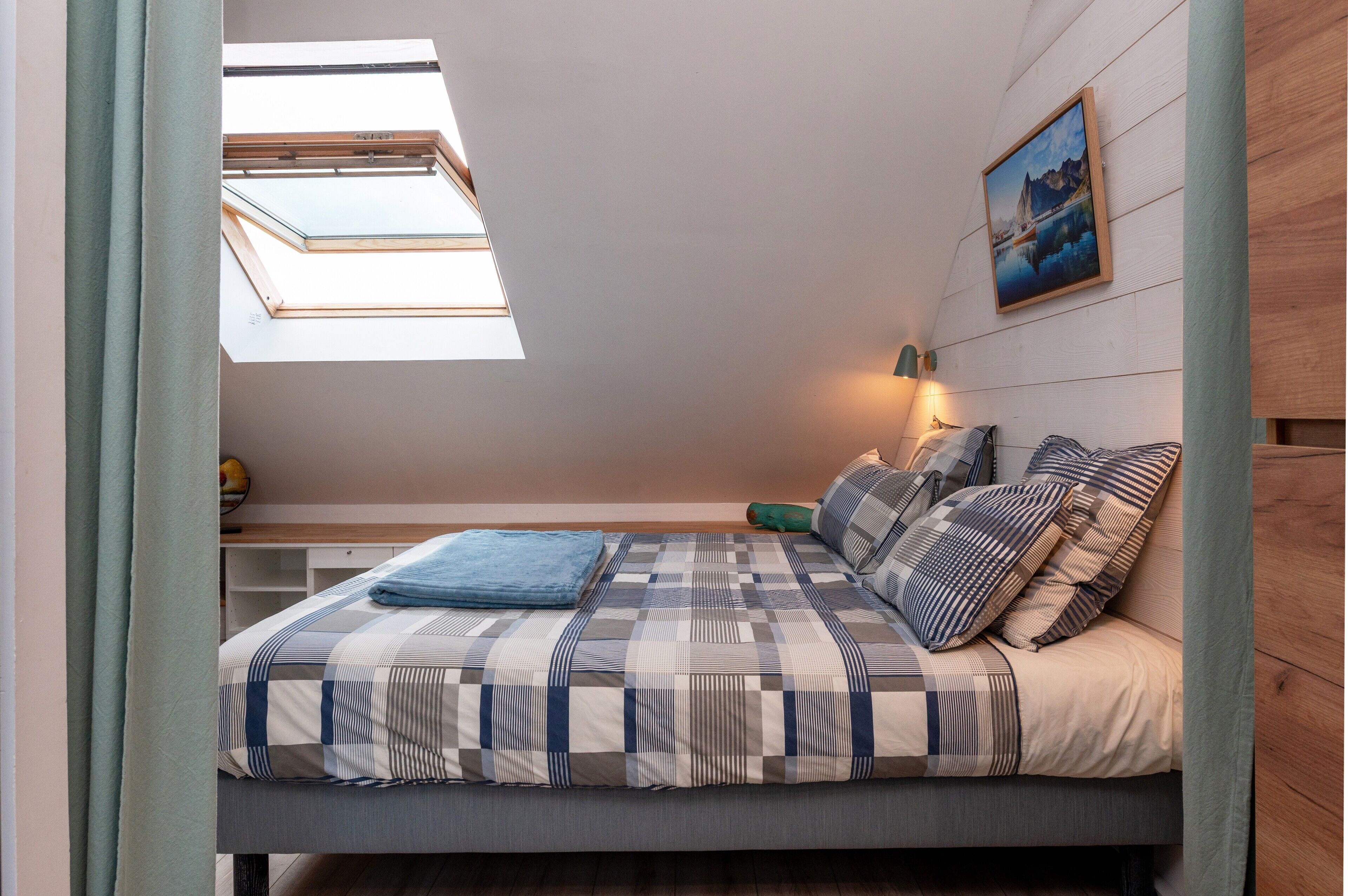 Ein Schlafzimmer mit Dachfenster, einem Bett mit gemusterten Betttextilien, einem Nachttisch, einer Lampe und einem Bild an der Wand.