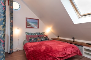 Une chambre à coucher avec une couvre-lit rouge, un tableau représentant une scène de plage et une lucarne.