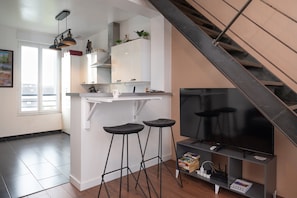 Une cuisine moderne dotée d’une cuisinière intégrée, de meubles blancs et d’un plan de travail noir. On y trouve deux tabourets de bar noirs et un téléviseur à écran plat posé sur un meuble gris.