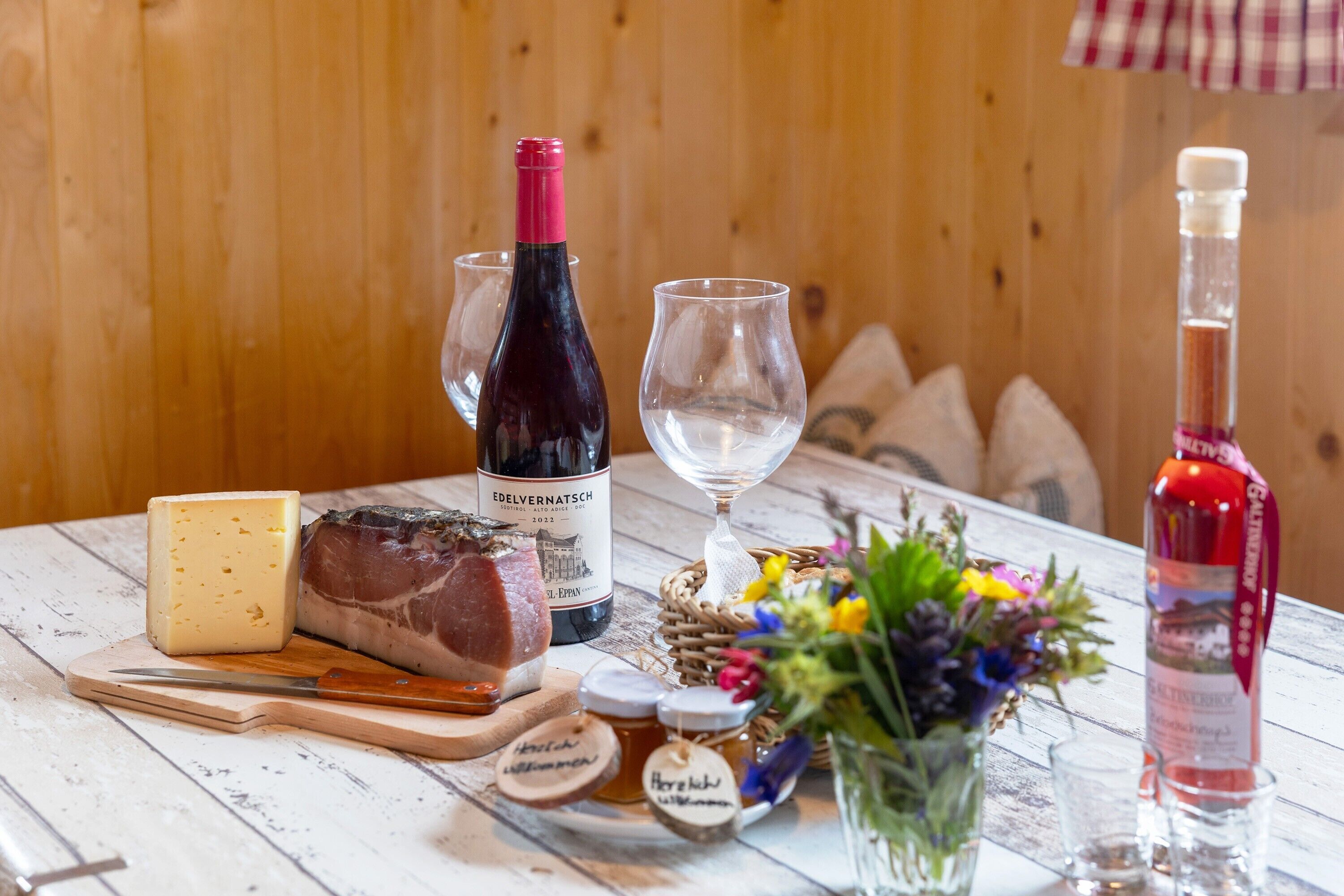 A wooden table with a bottle of wine, a glass, cheese, a loaf of bread, jars of honey, and a vase with flowers.