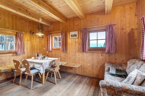 Interior de una cabaña de madera con una mesa de comedor, sillas, un sofá y una ventana con cortinas.
