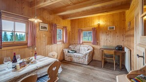 Interior d'una cabana de fusta amb una taula de menjador, cadires, un sofà amb flors i una taula petita amb un aparell de ràdio.