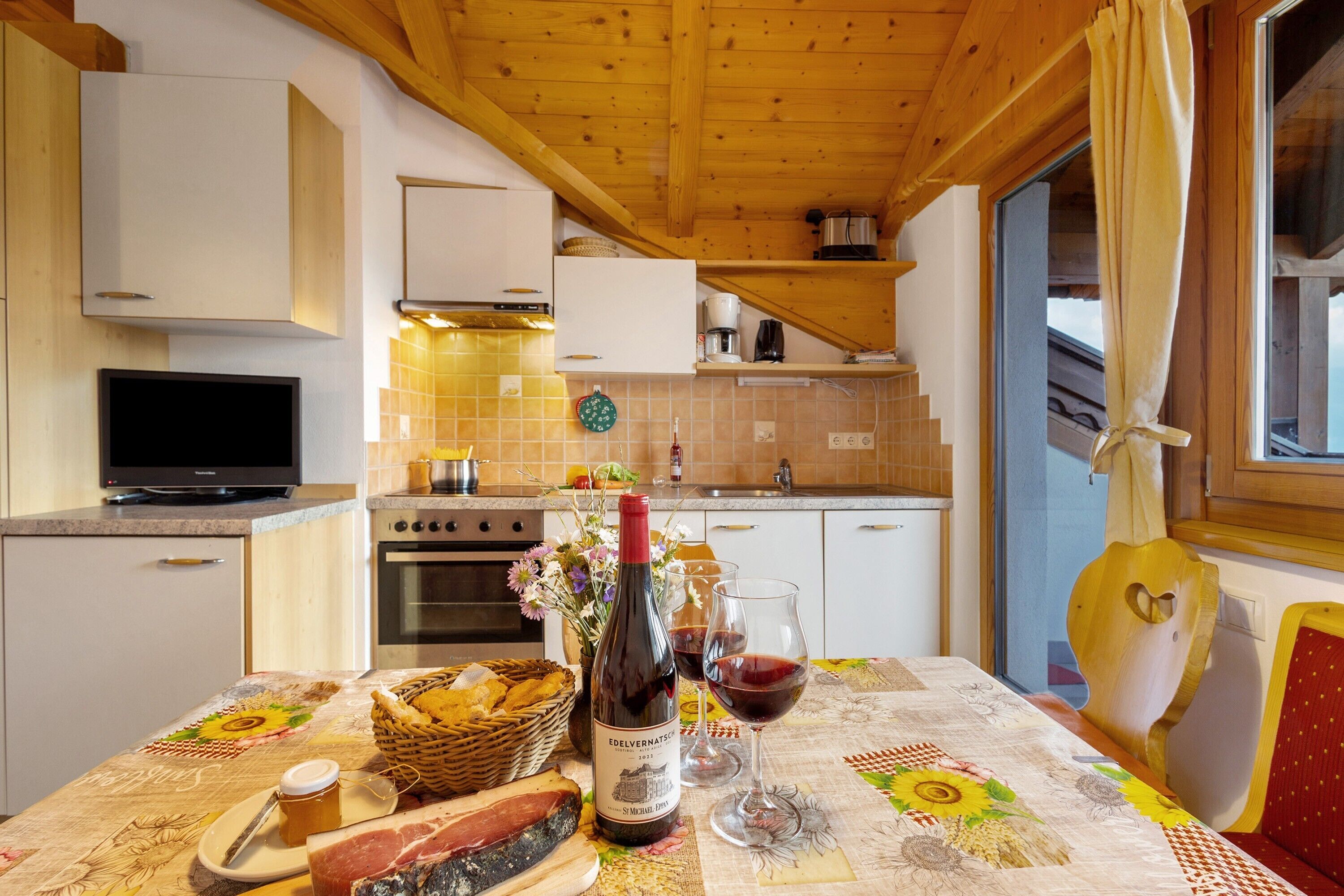 Une cuisine avec des meubles en bois, une télévision, une table à manger recouverte d’une nappe à motifs floraux et une bouteille de vin.