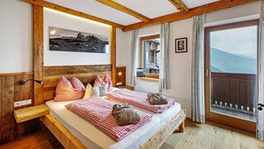 A wooden-framed bedroom with a bed, pillows, a bedside table, a wall-mounted lamp, a framed picture, a window with curtains, and a view of mountains. - Apartment 'Galtinerhof Almstadel' with Mountain View, Shared Terrace and Wi-Fi (Rodengo)