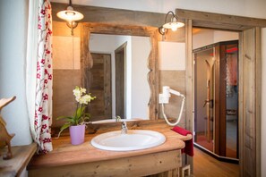 A bathroom with a wooden vanity, a white sink, a mirror with a wooden frame, a shower with a glass door, and a potted orchid.
