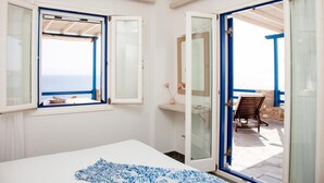 A bedroom with a bed, a window with a view of the sea, a bathroom with a mirror, and a chair outside.