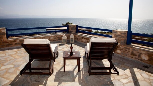 A terrace with stone flooring, two wooden lounge chairs, a table with a bottle and glasses, and a view of the sea.