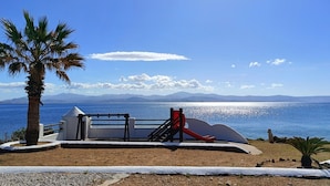 A playground with a slide and swings, palm trees, and a view of the sea and mountains.