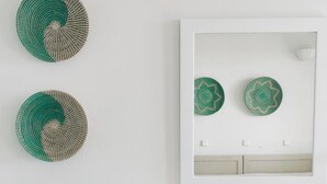 Ein Zimmer mit weißer Wand, zwei grünen gewebten Wandbehängen, einem spiegelnden, weiß umrahmten Spiegel, einem Regal mit einem Buch und einem abgenutzten Holzstuhl.