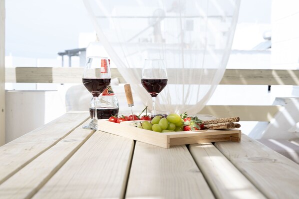Une table en bois sur laquelle sont disposés un plateau de raisins, des fraises et une bouteille de vin, le tout placé devant un fond blanc agrémenté d’un rideau transparent.