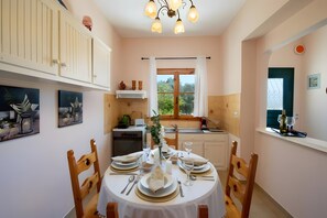A dining area with a round table set for six, wooden chairs, a white tablecloth, and a window with a view of greenery.