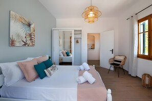 A bedroom with a bed, pillows, a bedside table, a hanging light fixture, a chair, and a view of a bathroom.