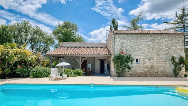 Un bâtiment en pierre avec un toit en tuiles, une piscine et un espace extérieur aménagé pour s’asseoir.