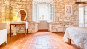 Une pièce aux murs de pierre, avec une fenêtre, un lit, une chaise et une petite table sur laquelle se trouvent une lampe et des fleurs.