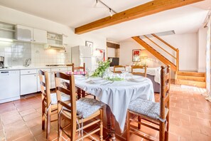 A dining area with a table set for four, wooden chairs, a kitchen in the background, and a staircase leading upstairs.