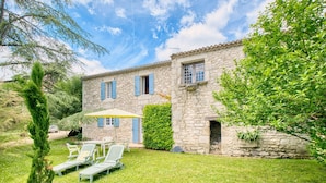 Une maison en pierre avec un jardin, une terrasse avec des chaises et une haie.