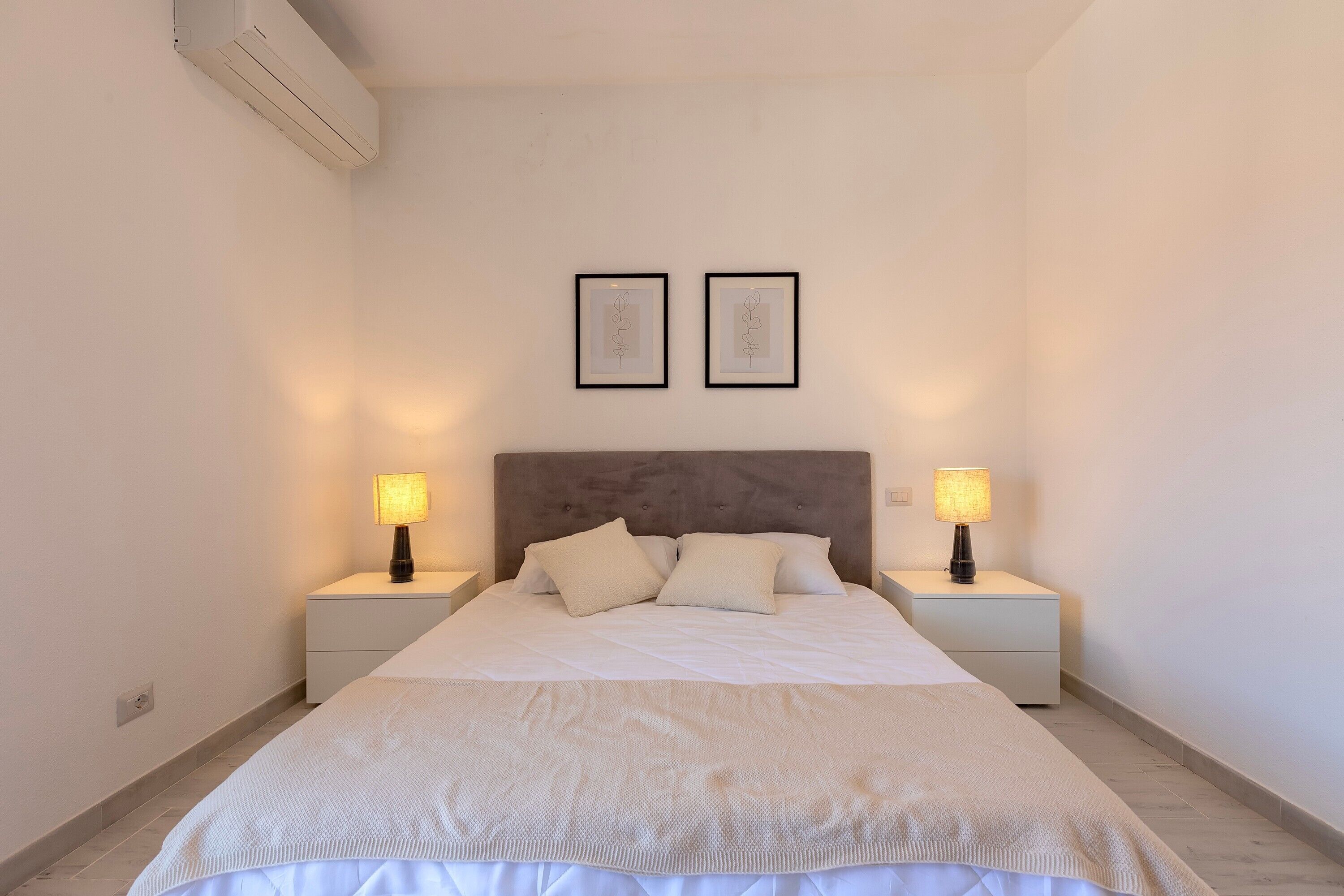 Un dormitorio con una cama, dos mesitas de noche, dos lámparas de pared y dos cuadros enmarcados.