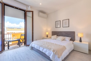 Et soveværelse med seng, sengeborde, lamper, et vindue med persienner og en balkon med bord og stole.