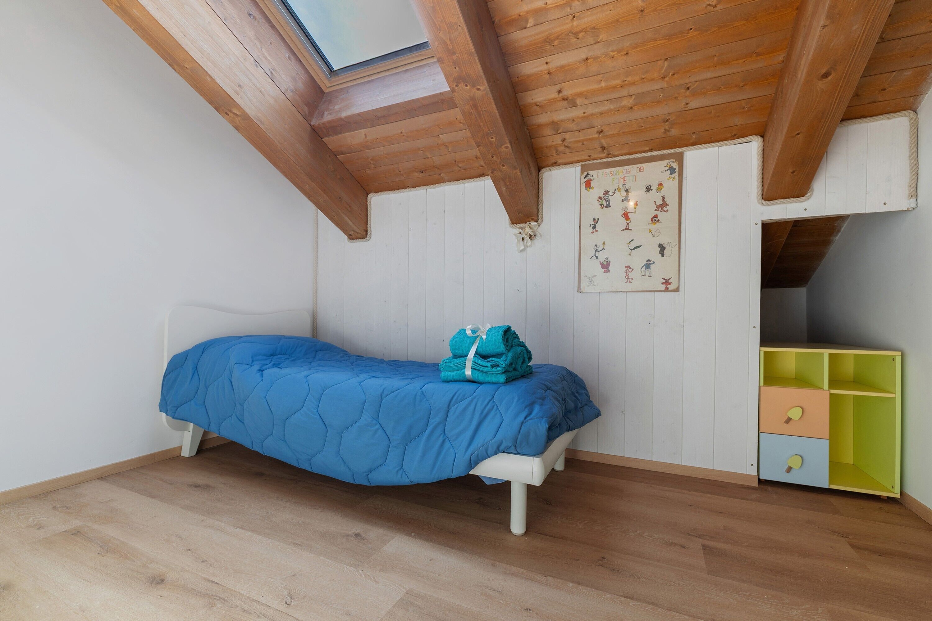 Una camera da letto con un letto, travi in legno al soffitto, un lucernario, una parete con un'opera d'arte per bambini e un contenitore colorato.