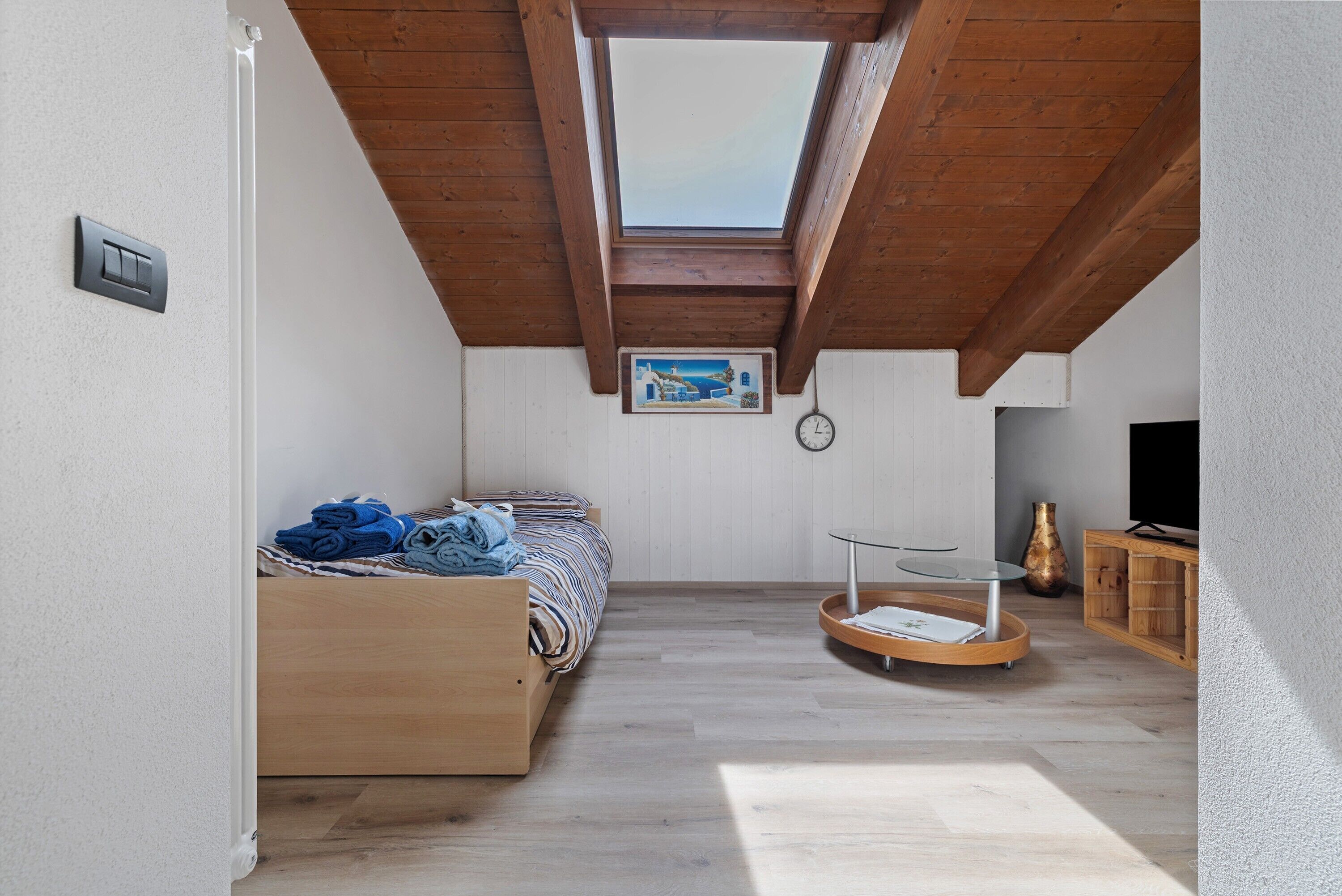 A room with a sloped ceiling, a bed, a small table, and a painting on the wall.