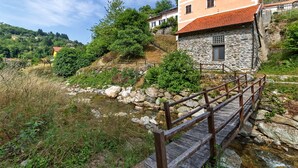 Un edificio de piedra con un puente de madera sobre un arroyo, rodeado de vegetación y colinas.