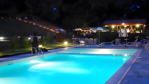 A swimming pool with a clear edge, a covered seating area with umbrellas, and a building with lights in the background. - Apartment 'Malva' with Shared Pool, Wi-Fi and Air Conditioning (Livorno)