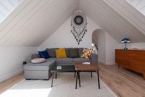 A loft-style room with a gray sectional sofa, a wooden coffee table, a dreamcatcher, a dresser, and a lamp.