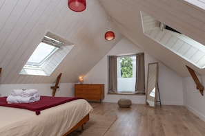 A bedroom with a sloped ceiling, skylight windows, a bed with a red blanket, a wooden dresser, and a round ottoman.