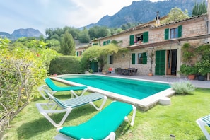 Ein Pool mit Liegestühlen, ein Steinhaus mit grünen Fensterläden und Berge im Hintergrund.