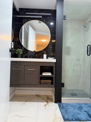 A bathroom with a marble vanity, a round mirror, a shower with a glass door, and a blue rug. - THE GUEST HOUSE WEST EDMONTON MALL (Edmonton)