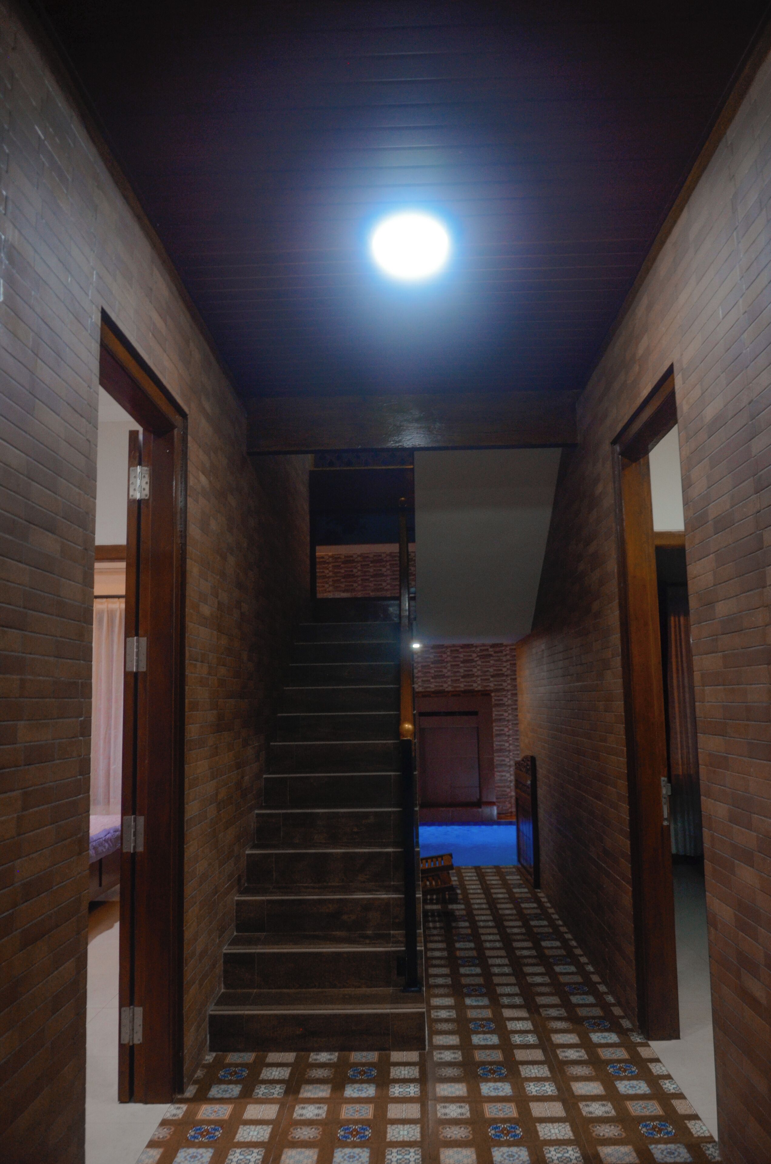 A narrow hallway with a staircase leading to a landing. The walls are made of brick, and the floor has patterned tiles.
