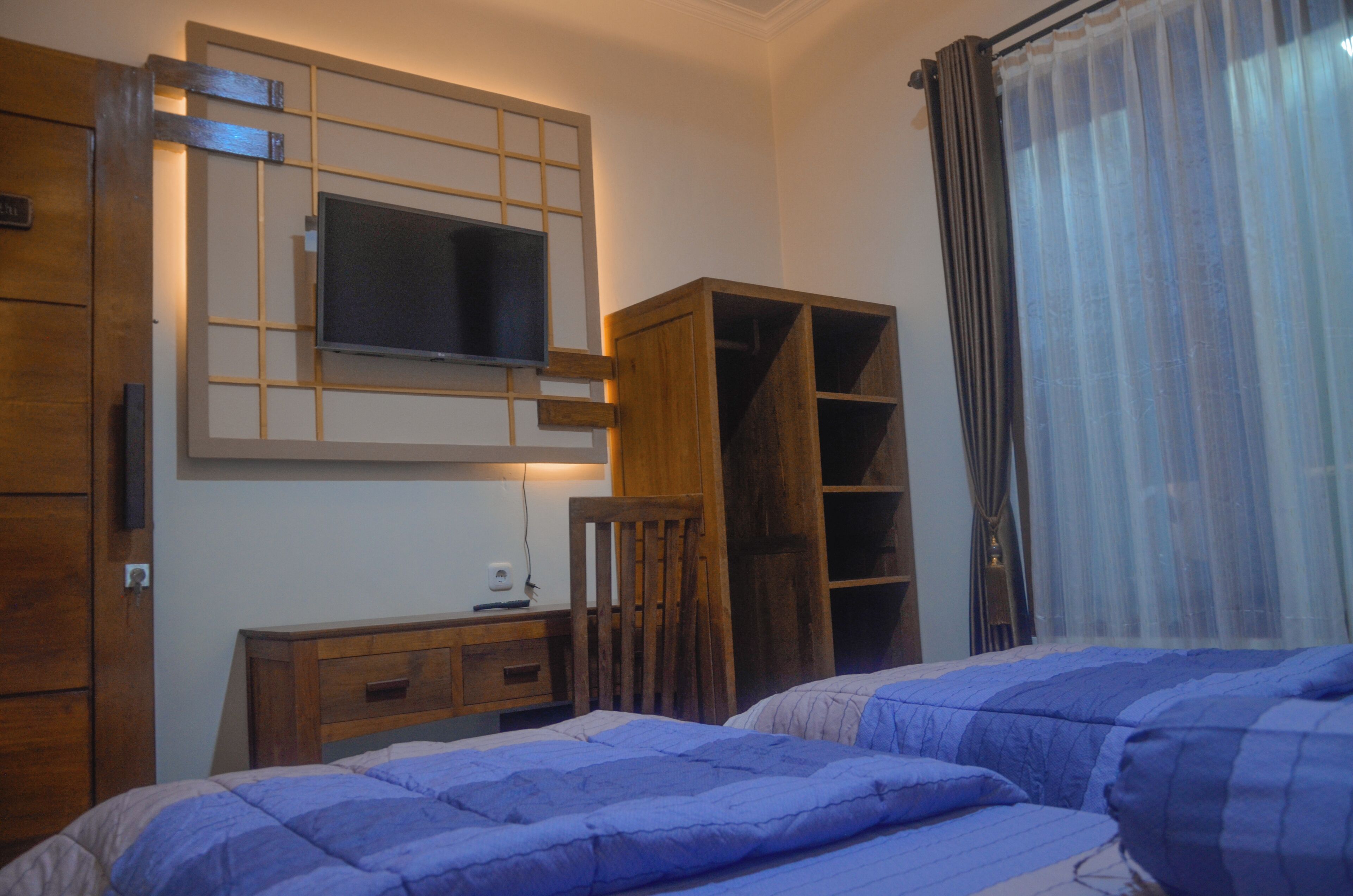 A bedroom with two beds, a TV mounted on the wall, wooden furniture, and a window with curtains.