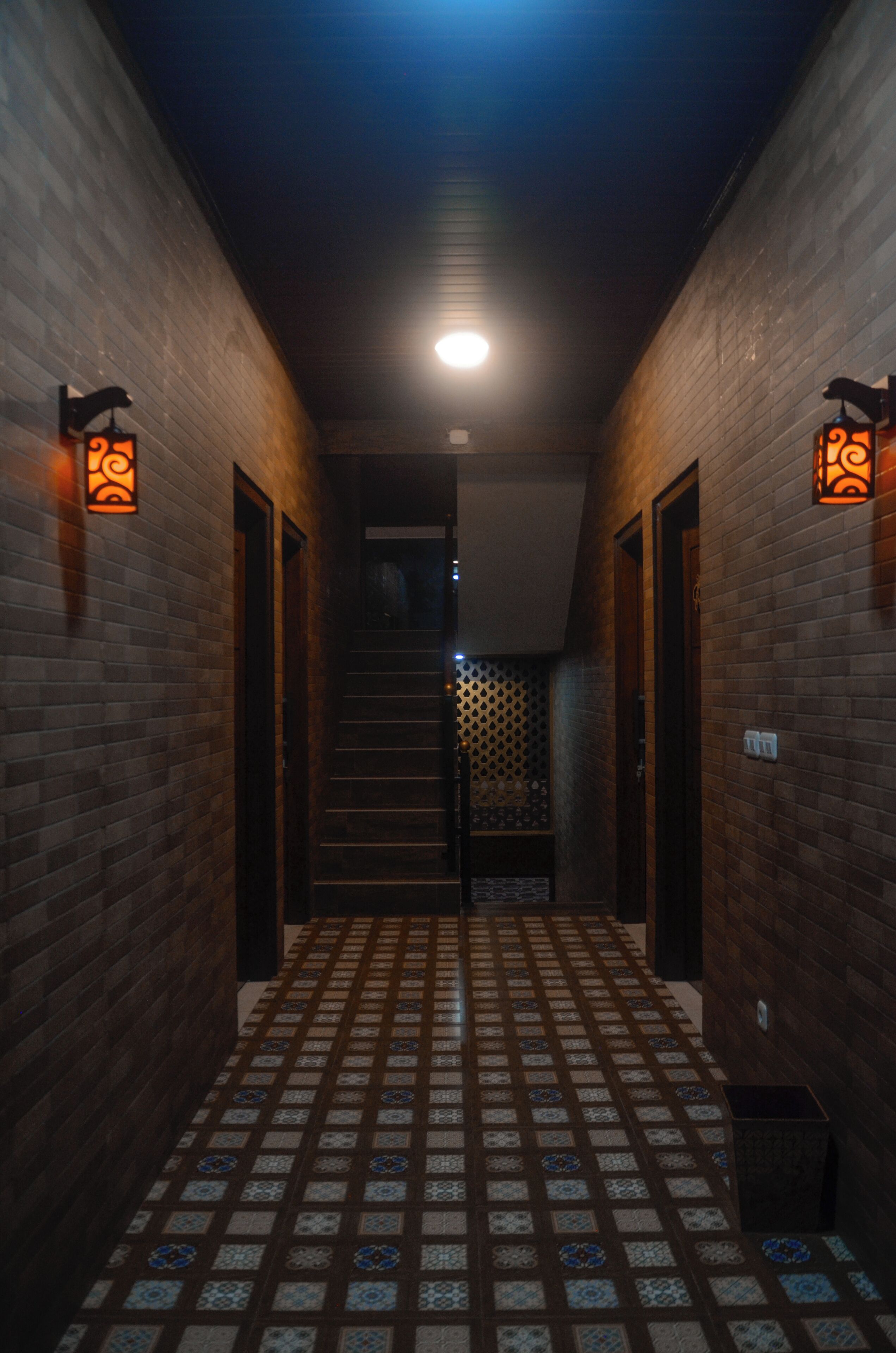 A narrow hallway with patterned tile flooring, brick walls, and two wall-mounted light fixtures.