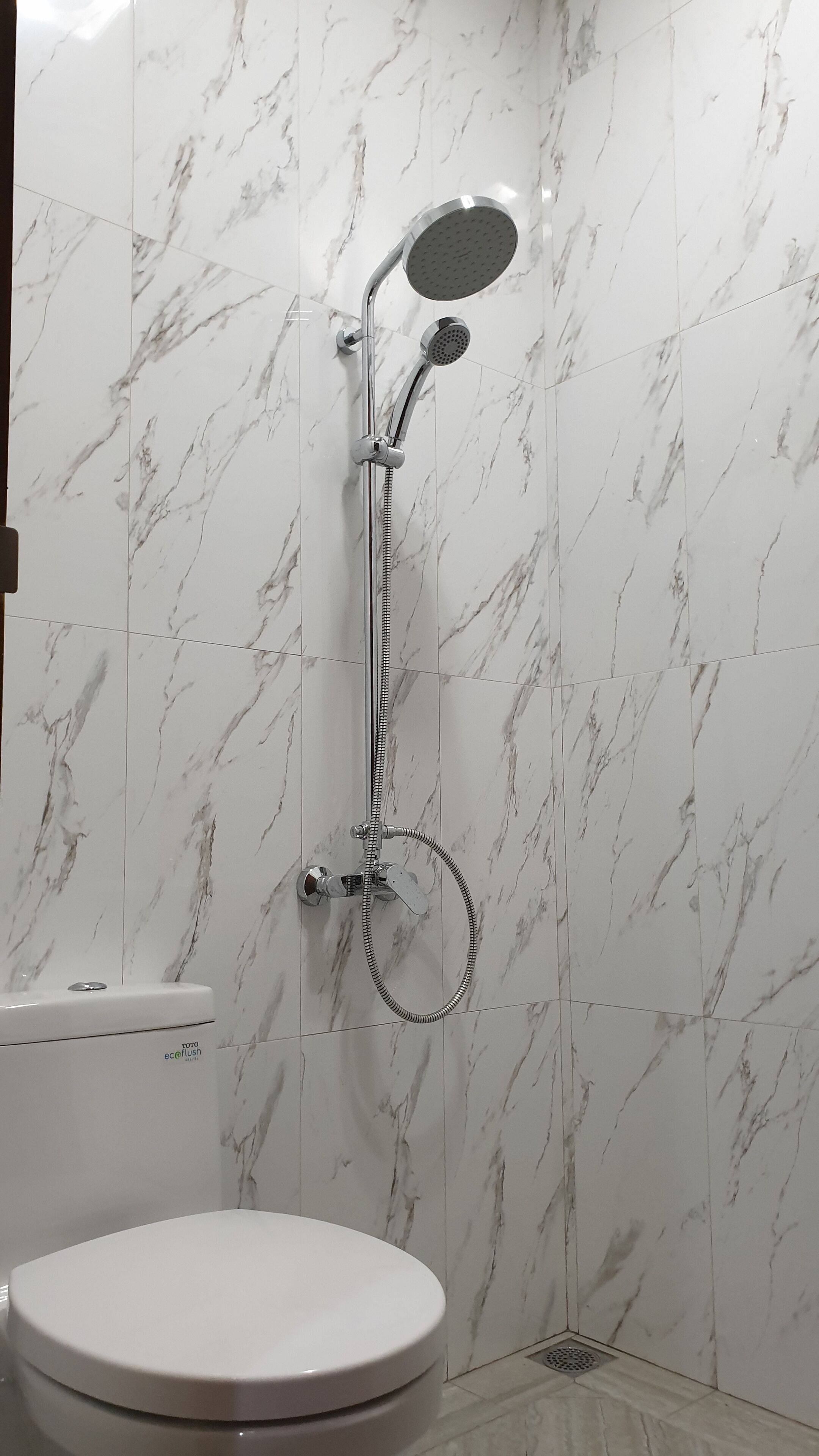 Une salle de bain avec des toilettes blanches, une douche équipée d’un pommeau de douche à main et un mur au motif de marbre.