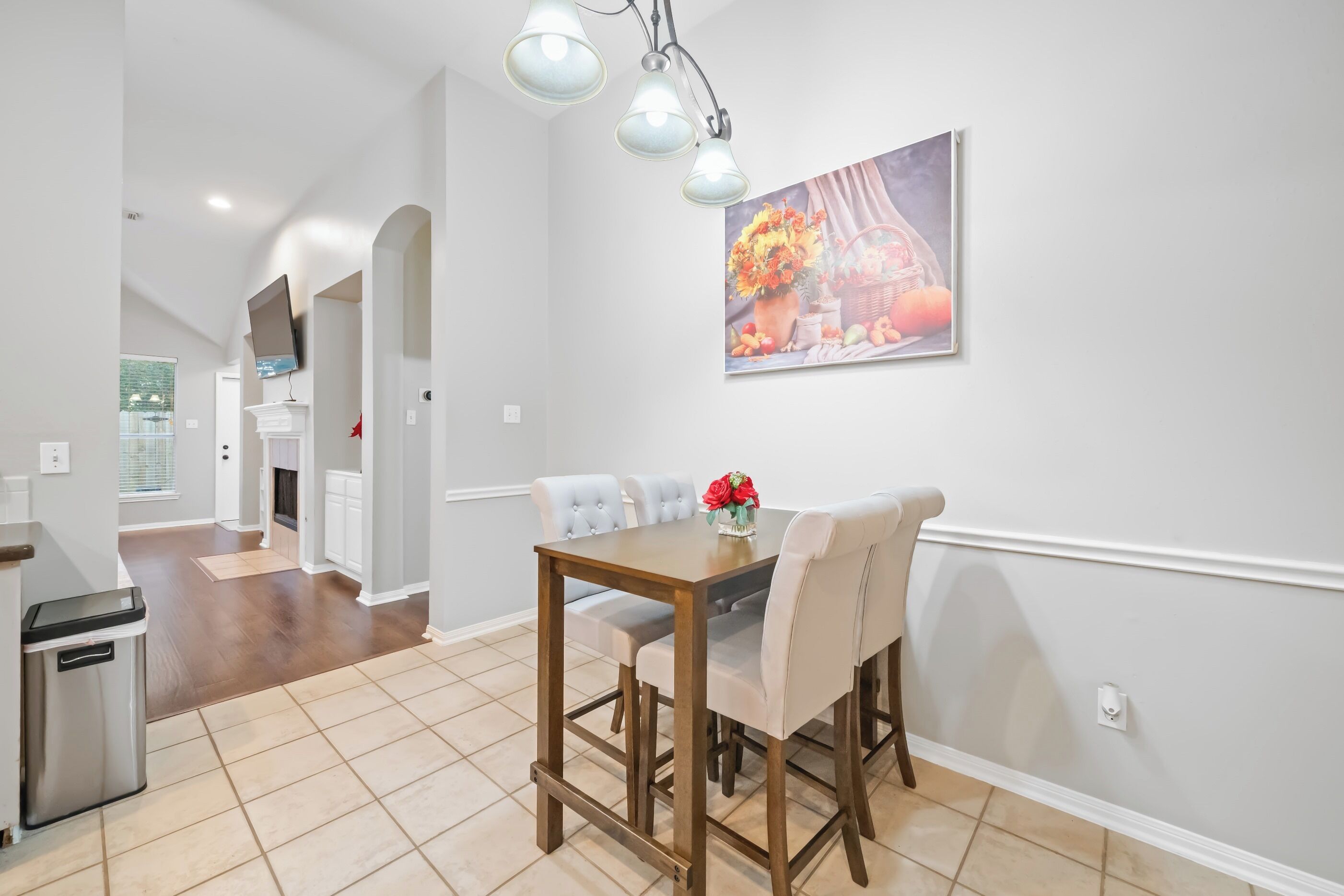 A dining area with a wooden table and chairs, a painting on the wall, and a trash bin.