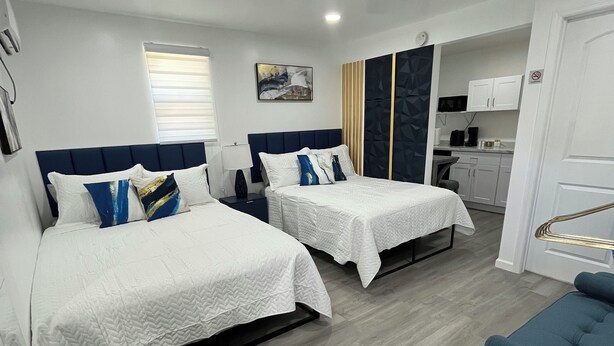 A hotel room with two beds, a blue armchair, a small round ottoman, a kitchenette, and a bathroom. - Cozy Private Tiny Guest House (Jacksonville)