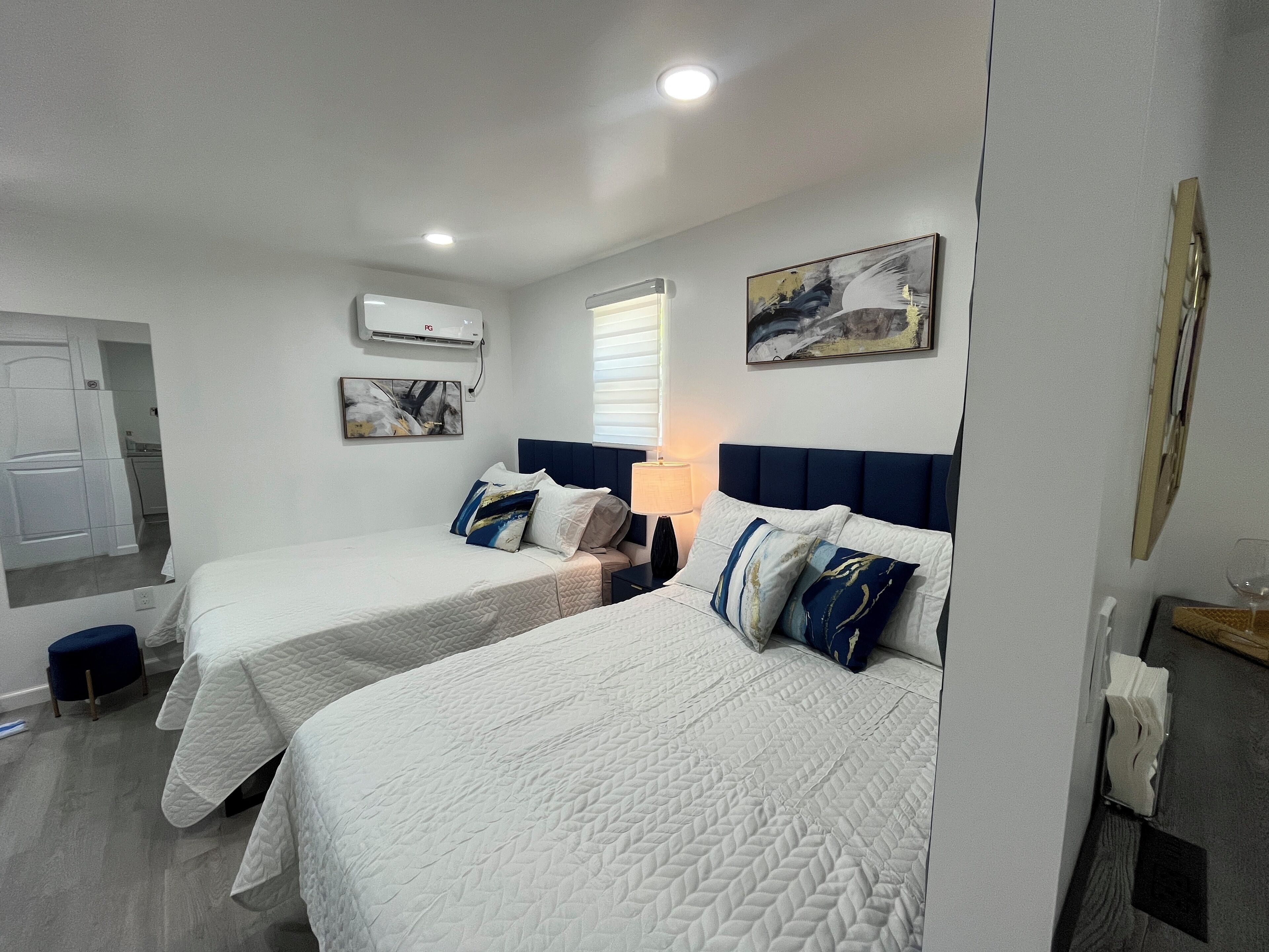 A hotel room with two beds, a nightstand, a lamp, a painting, and a window with blinds.